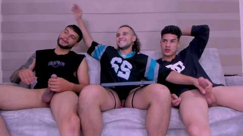 Media: Video of three shirtless, erect men with varying body types and ethnicities, sitting on a white couch, in a modern, minimalist room.