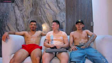 Media: Video of three shirtless Asian men in casual attire, sitting on a couch in a modern living room with a marble wall.