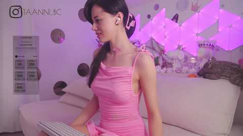 Media: Video of a young woman with light skin and long dark hair, wearing a pink dress, typing on a keyboard in a modern, softly lit room with purple LED lights and a white couch.