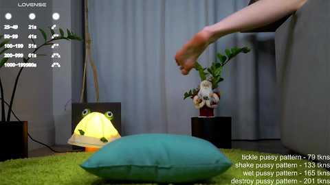 Media: A video of a person interacting with a toy frog lamp and a green pillow on a green carpet, surrounded by potted plants and a monitor displaying a 3D model.