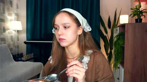 Media: Video of a young woman with long brown hair, wearing a white headscarf and brown dress with lace details, eating soup in a cozy, dimly lit living room with teal curtains, wooden furniture, and potted plants.