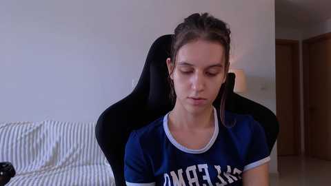 Media: A video of a young woman with light skin and braided hair, wearing a navy blue t-shirt, sitting on a black gaming chair in a minimalist room with a striped sofa and wooden door in the background.