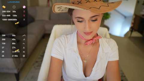 Media: Video of a young woman with light skin, wearing a white blouse and a large, straw sombrero, sitting indoors; she has a pink and white striped neckerchief and a gold necklace.