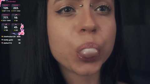 Media: A close-up video of a young woman with fair skin, long black hair, and pierced nose, making a pouty face while holding a lollipop, against a dark background.