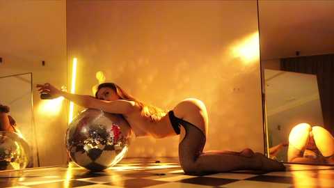 Media: Video of a topless woman with long brown hair, wearing black lingerie and fishnet stockings, kneeling on a checkered floor, hugging a glittery disco ball, in a dimly lit, modern room with mirrored walls.