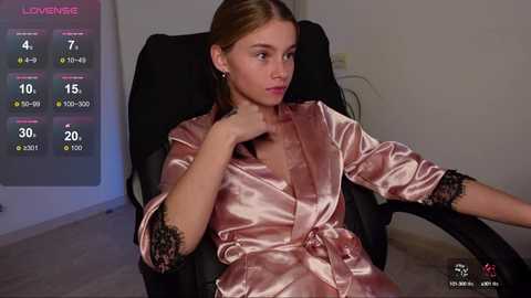 Media: A video of a young Caucasian woman with light skin, brown hair, and light makeup, wearing a silky, pink satin robe, seated in a black office chair, with a digital timer on a wall behind her.