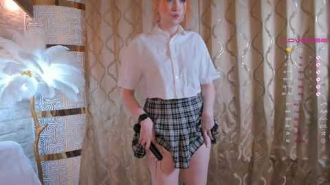 Media: Video of a pale-skinned, slim, androgynous person with shoulder-length blonde hair, wearing a white button-up shirt, black plaid skirt, and black wristbands, standing in front of gold-patterned curtains and a white floral arrangement.