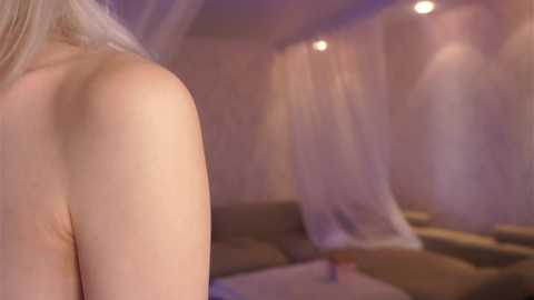 Media: A video showing a nude woman with fair skin and long blonde hair, viewed from the back, standing in a softly lit room with sheer curtains and a gray couch.