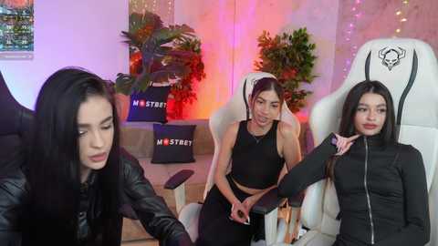 Media: Video of three women in a gaming room: one with long black hair in a leather jacket, another in a black crop top, and a third in a black jacket, seated in a white gaming chair.