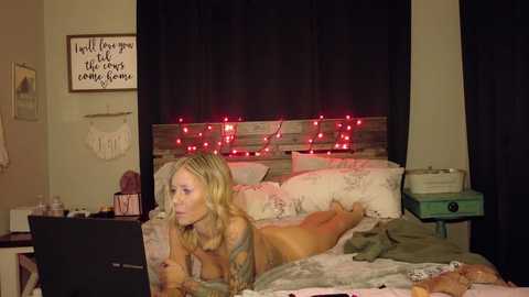 Media: A video of a blonde, tattooed woman lying naked on a bed with a floral duvet, surrounded by string lights, in a cozy, dimly-lit bedroom.