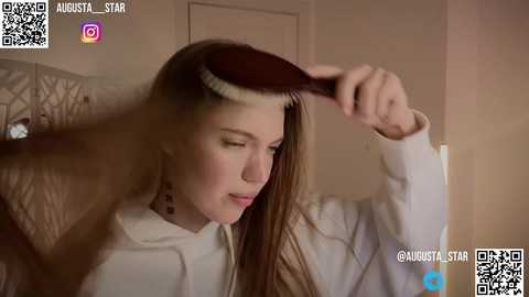 Media: Video of a young woman with long brown hair, wearing a white hoodie, brushing a wig with a dark brown ponytail in a dimly lit room.