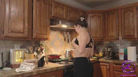 Media: Video of a woman in a kitchen, wearing a black bra and black pants, cooking. Wooden cabinets, utensils, and appliances fill the background.