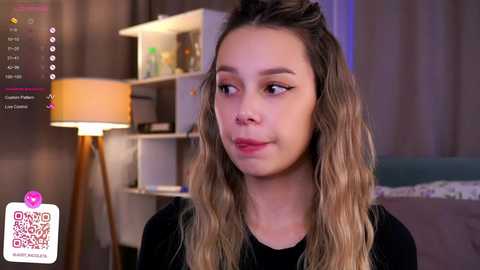 Media: Video of a young woman with long, wavy blonde hair, wearing a black top, sitting in a modern, dimly lit room with a white shelf and lamp.