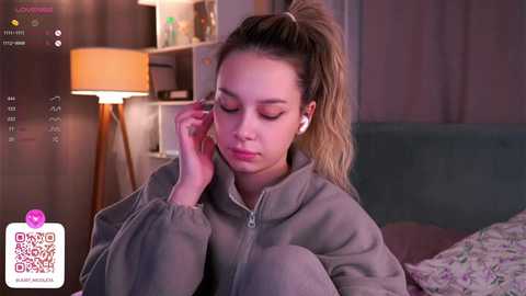 Media: Video of a young woman with fair skin, brown hair tied in a ponytail, wearing headphones, and a gray hoodie, sitting on a bed in a dimly lit room with a lamp and a bookshelf in the background.