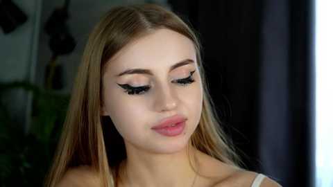 Media: Video of a young Caucasian woman with long, straight blonde hair and fair skin, wearing black eyeliner and pink lipstick, indoors with dark curtains in the background.