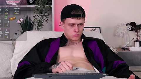 Media: Video of a shirtless, fit young man with tattoos, wearing a black and purple jacket, black cap, and headphones, focused on a laptop, in a modern living room with a white sofa, plant, and decorative items.