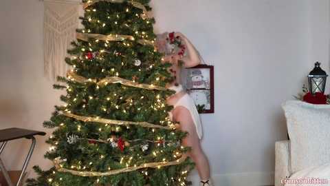 Media: Video of a white woman in a white tank top, wrapping a gold ribbon around a decorated Christmas tree. Background features a beige wall, a macrame hanging, and a white couch.