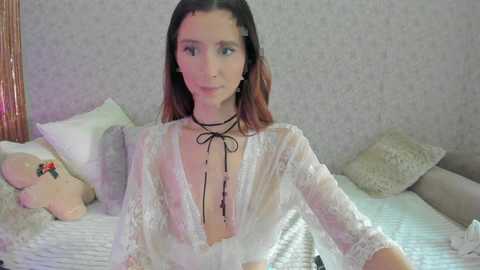 Media: A video of a fair-skinned woman with shoulder-length dark hair, wearing a sheer lace robe with a deep V-neckline, sitting on a bed with plush pillows and a teddy bear.