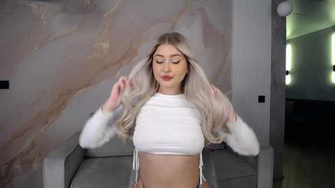 Media: Video of a fair-skinned, blonde woman with long, wavy hair, wearing a white, fuzzy crop top and matching pants, standing in a modern living room with marble walls and a grey sofa.