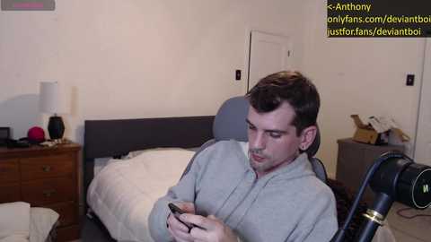 Media: Video of a man with short brown hair, wearing a grey hoodie, sitting on a bed in a modest bedroom, holding a phone, with a camera tripod visible.