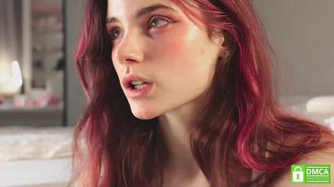 Media: Video of a young woman with fair skin and long, wavy red hair, gazing intently. She has subtle blush on her cheeks, and the background shows a blurred, modern bedroom with a mirror.