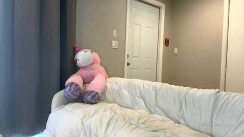 Media: Video of a plush pink unicorn toy sitting on a white quilted sofa in a beige-walled room with a closed white door and light switches.