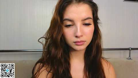 Media: Video of a young woman with long, wavy brown hair, light skin, and delicate facial features, standing indoors against a plain white wall, wearing a black top.