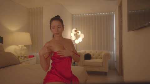 Media: Video of an Asian woman in a red satin dress, covering her breasts, in a dimly lit, modern bedroom with beige walls, a white couch, and a lamp.