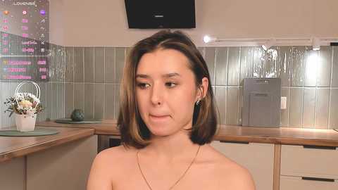 Media: Video of a young woman with straight, shoulder-length brown hair, wearing a gold necklace, standing in a modern kitchen with tiled backsplash, countertop, and appliances.