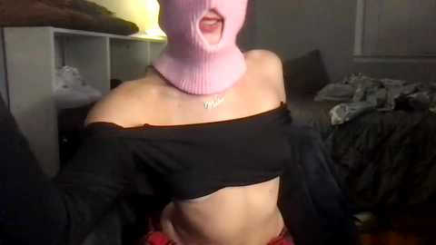 Media: Video of a woman with a pink knitted hat covering her face, wearing a black off-shoulder crop top revealing her midriff, and red shorts, standing in a dimly lit bedroom.