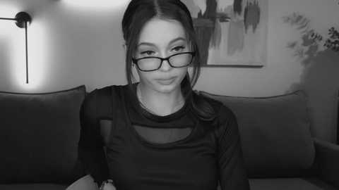 Media: A black-and-white video of a young woman with glasses, wearing a dark, long-sleeve top, seated on a sofa. The background features a minimalist painting and a modern light fixture.