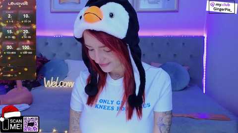 Media: Video of a pale-skinned woman with red hair wearing a white t-shirt and a penguin hat, smiling in a dimly lit bedroom with a bed and a TV displaying a chat window.