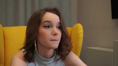 Video of a young woman with shoulder-length brown hair, wearing a sleeveless light grey top and silver earrings, sitting on a bright yellow armchair, against a grey curtain backdrop.