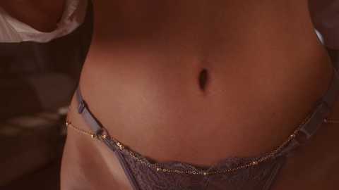 Media: Video of a light-skinned woman's lower abdomen and hip, wearing blue lace panties with a gold chain belt, in a dimly lit, indoor setting.