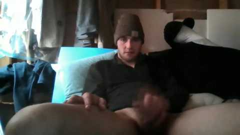 Media: Video of a young man with a beard in a beanie, wearing a dark shirt, lying on a bed with a black dog. Background includes a blue blanket, a jacket, and a wooden door.