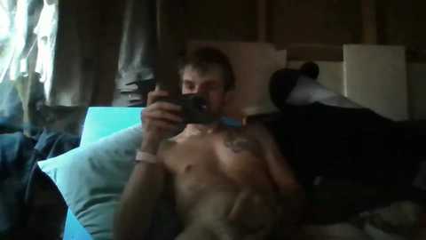 Media: A video of a shirtless man with a tattoo on his chest, holding a phone, lying on a bed with a dog. Background shows a cluttered room with clothes, a blue pillow, and a wooden cabinet.