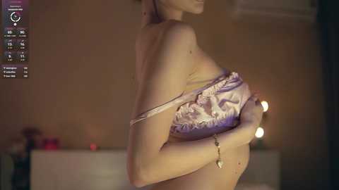 Video of a fair-skinned woman with medium-sized breasts, partially topless, holding a light-colored bra with a gold pendant. Background shows a dimly lit, beige room with a blurred bed and a lamp.