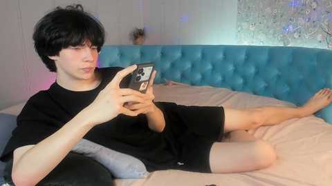 Media: Video of a young man with short black hair, pale skin, and a slim build, wearing a black t-shirt and shorts, lying on a bed with a turquoise tufted headboard, holding a smartphone, in a brightly lit room.