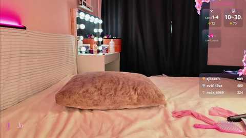 Media: Video of a cozy bedroom with a beige cushion on a white bedspread, pink and black lingerie on the floor, and a vanity mirror reflecting a lit-up room.