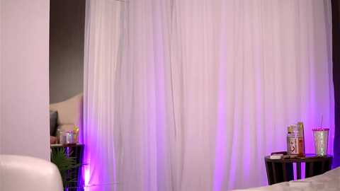 Media: Video of a serene, minimalist room with white curtains, purple lighting, a black side table holding a candle and book, and a white leather couch in the foreground.