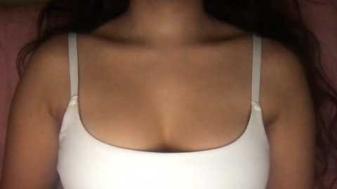 Media: A video of a woman's upper torso in a white, thin-strapped tank top, with long, dark hair cascading over her shoulders. The background is a soft pink.