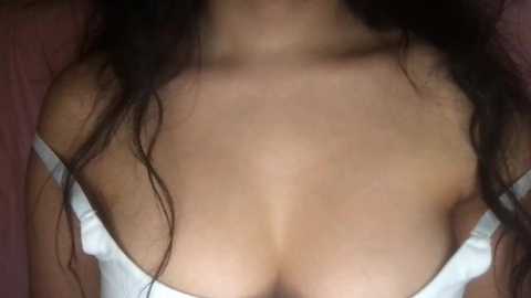 Media: A video showing the upper body of a woman with long, dark hair and light brown skin, wearing a white bra, highlighting her large, round breasts. The background is a blurred, dark fabric.