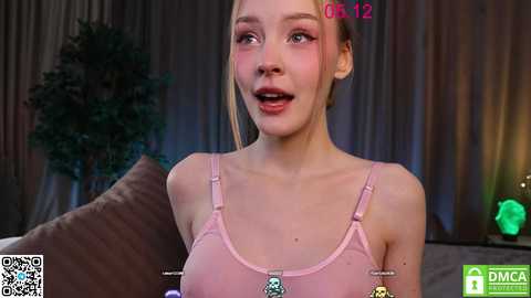 Media: Video of a young woman with fair skin, light brown hair, and green eyes, wearing a pink spaghetti-strap tank top, smiling in a cozy, dimly lit living room.