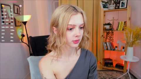 Media: Video of a young blonde woman with fair skin, wearing a gray off-shoulder top, seated indoors. Background includes a bookshelf, a lamp, and a table.