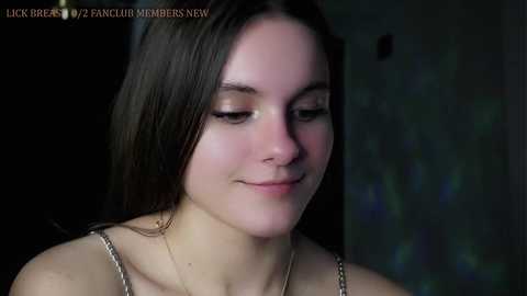 Media: Video of a young, fair-skinned woman with long, dark hair, wearing a thin strap top, softly smiling. Background is dark and blurry, with text \"LICK BREAST 2 PICNIO MEMBERS NOW\" in the top left corner.