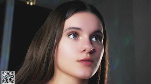 Media: Video of a young woman with long brown hair, fair skin, and blue eyes, wearing a small nose ring, looking pensive.