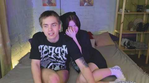 Media: Video of two young men on a bed in a dimly lit room. One is shirtless, wearing shorts and a t-shirt; the other wears a black hoodie, hiding his face.