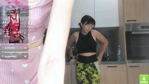 Media: Video of an Asian woman in a black sports bra and green leggings, standing in a modern kitchen, with a smartphone video recording herself.