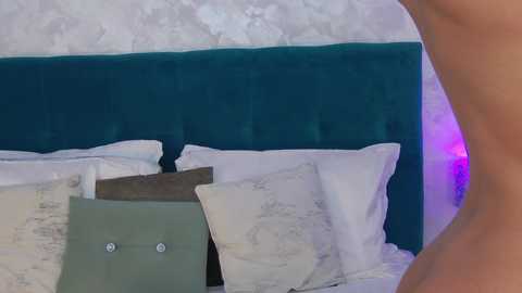 Media: Video of a nude man standing beside a bed with a teal velvet headboard, white pillows, and a grey pillowcase. Background features a textured white wall with purple lighting.