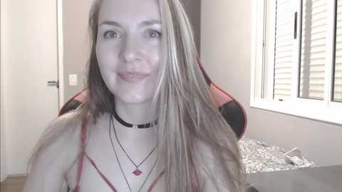 Media: Video of a fair-skinned, blonde-haired woman with blue eyes, wearing a black choker and multiple necklaces. She smiles in a brightly lit room with white shutters and a red and black gaming chair.